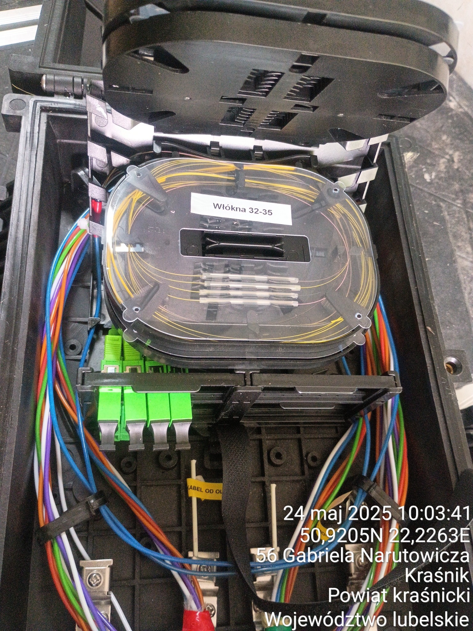 Otwarta skrzynka telekomunikacyjna z widocznymi światłowodami, złączami i okablowaniem. Wewnątrz etykieta 'Włókna 32-35'.