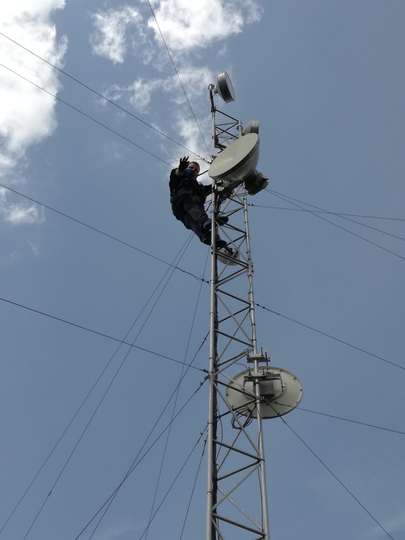 Pracownik na szczycie wieży telekomunikacyjnej, instaluje antenę satelitarną na tle błękitnego nieba z chmurami. Ujęcie z dołu podkreśla wysokość konstrukcji.