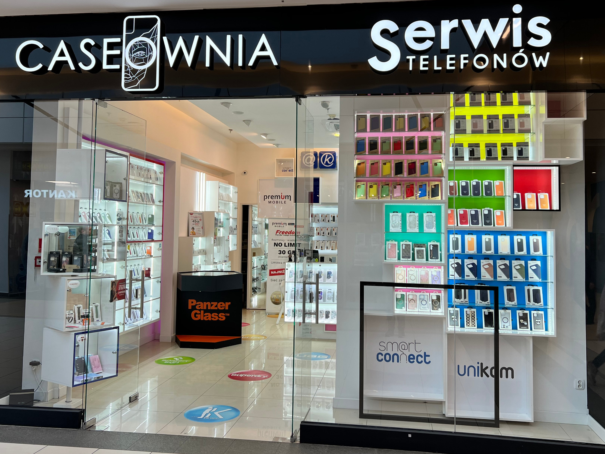 Wejście do sklepu z akcesoriami do telefonów Caseownia Serwis Telefonów, widoczne liczne ekspozycje z etui na telefony, szkła ochronne i inne akcesoria. Na podłodze naklejki z logo różnych marek.