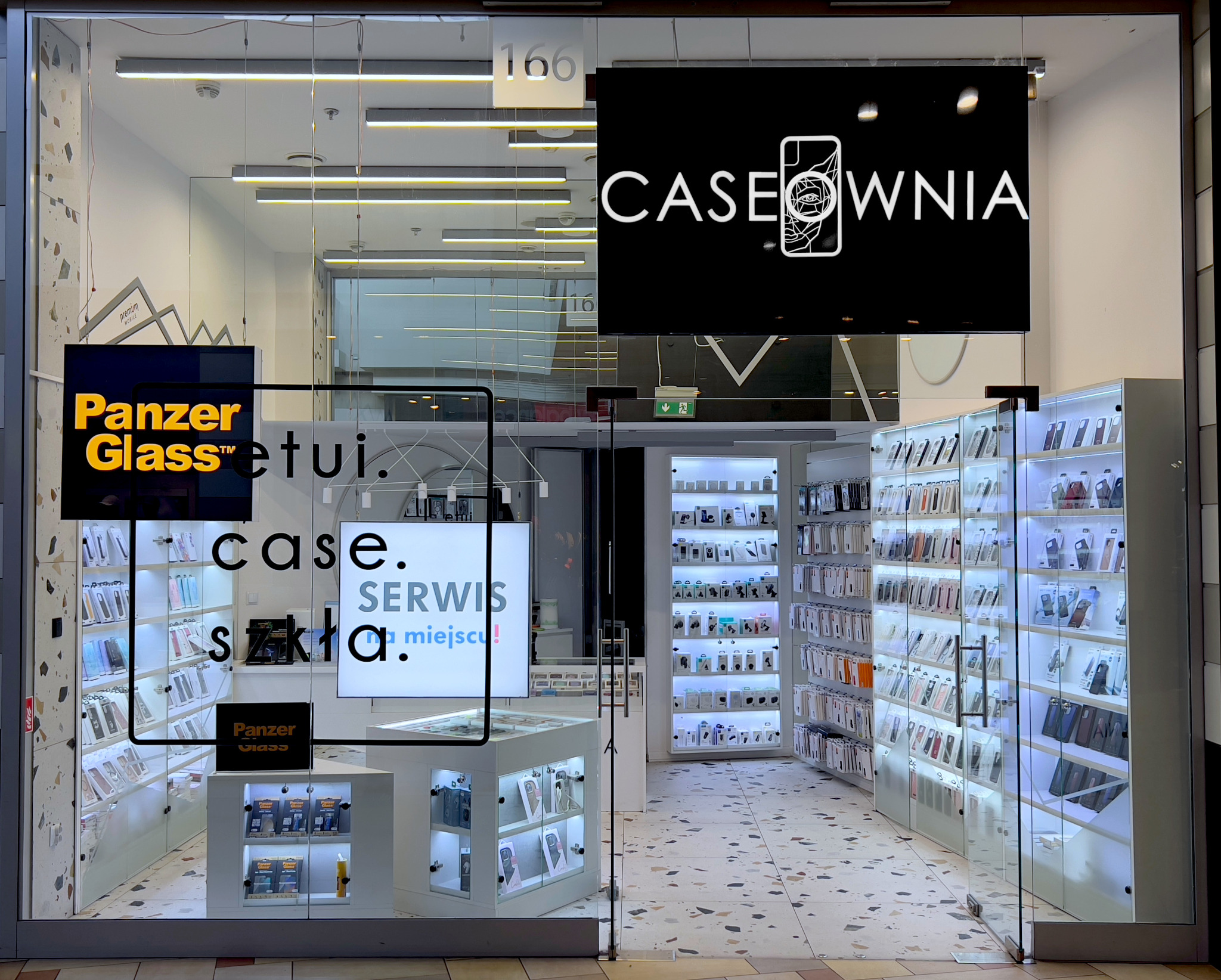 Wejście do sklepu z akcesoriami do telefonów, Caseownia, z widocznymi gablotami wypełnionymi etui, szkłami ochronnymi i innymi akcesoriami. Na szybie naklejki z logo Panzer Glass i informacją...