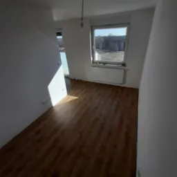 Pusty pokój po remoncie z nowymi panelami podłogowymi, świeżo pomalowanymi ścianami na biało, grzejnikiem pod oknem i wystającym kablem od lampy.