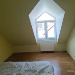 Pokój na poddaszu z oknem w kształcie trapezu, świeżo pomalowane ściany w odcieniach żółci, parkiet ułożony w jodełkę, widoczny fragment łóżka z pikowaną narzutą.