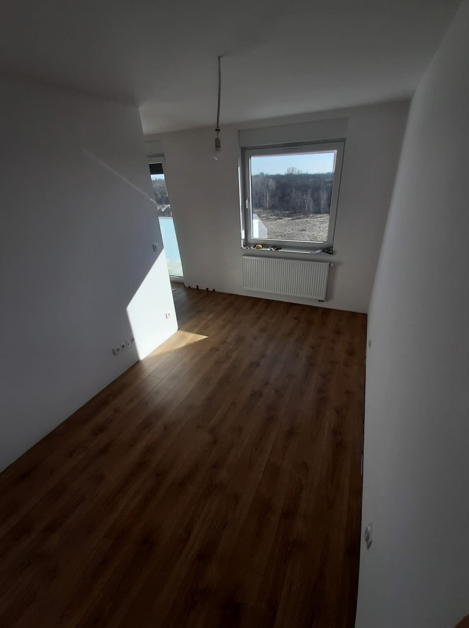 Pusty pokój po remoncie z nowymi panelami podłogowymi, świeżo pomalowanymi ścianami na biało, grzejnikiem pod oknem i wystającym kablem od lampy.