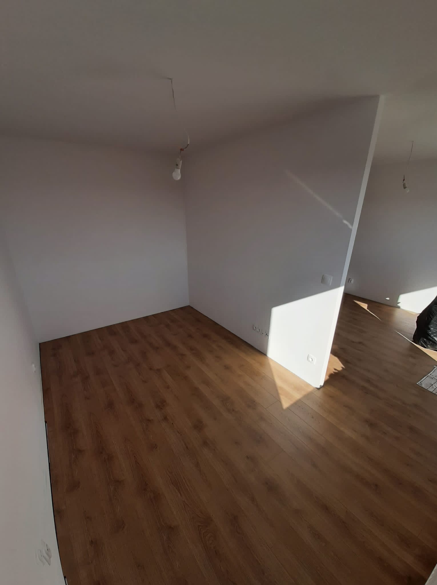 Pusty pokój z ułożonymi panelami podłogowymi w odcieniach brązu, świeżo pomalowanymi białymi ścianami i widocznym okablowaniem oświetleniowym.