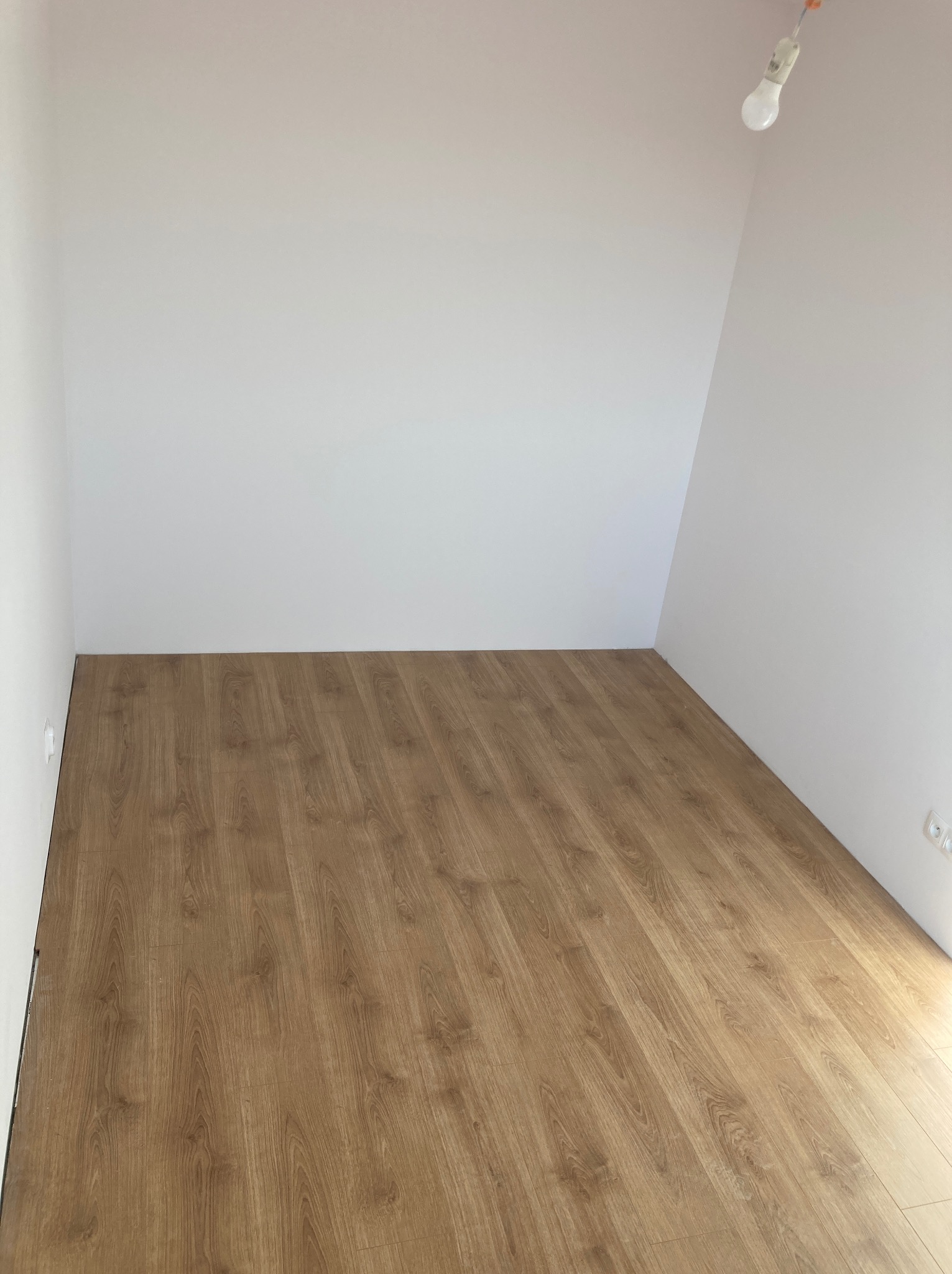 Ukończone układanie paneli podłogowych w pustym, jasnym pokoju z białymi ścianami, widoczny surowy kabel oświetleniowy.