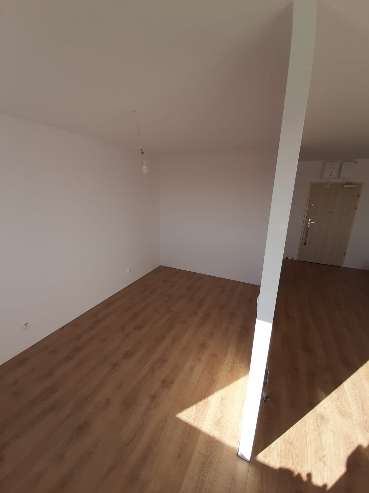 Pusty pokój z nową drewnianą podłogą, świeżo pomalowanymi białymi ścianami i wystającą kolumną, oświetlony naturalnym światłem wpadającym przez drzwi.