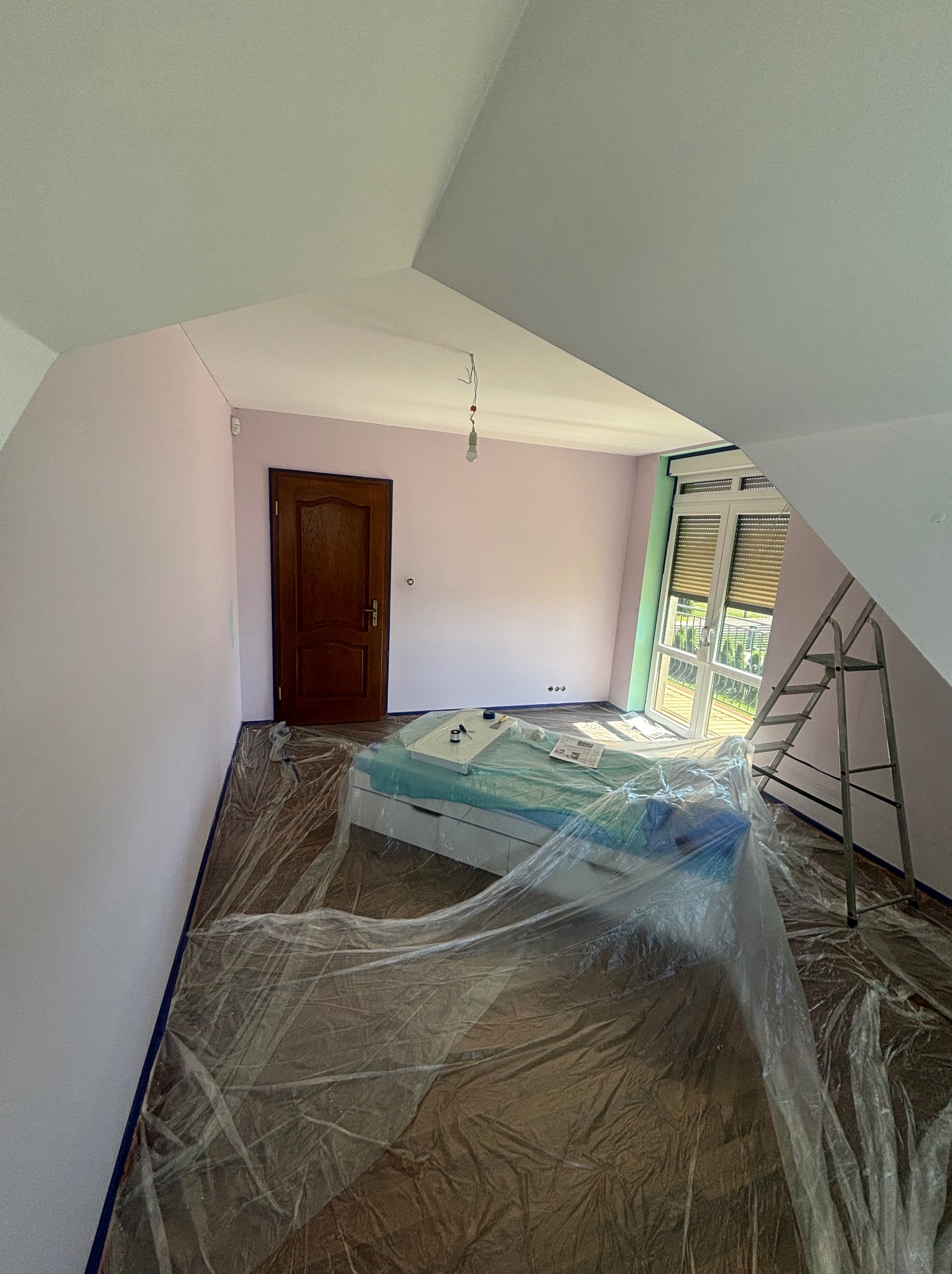 Pokój na poddaszu w trakcie odświeżania ścian na delikatny róż, podłoga zabezpieczona folią malarską, drabina oparta o ścianę przy oknie.