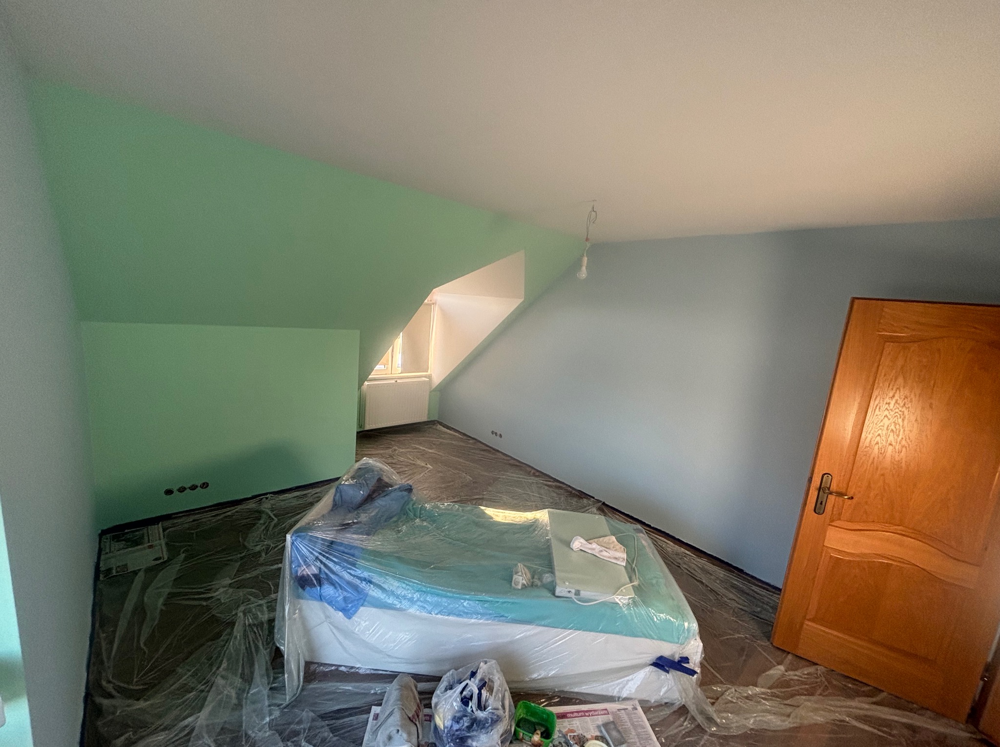 Pokój na poddaszu w trakcie malowania: ściany częściowo pomalowane na zielono i niebiesko, meble zabezpieczone folią malarską, widoczne gniazdka elektryczne i drewniane drzwi.