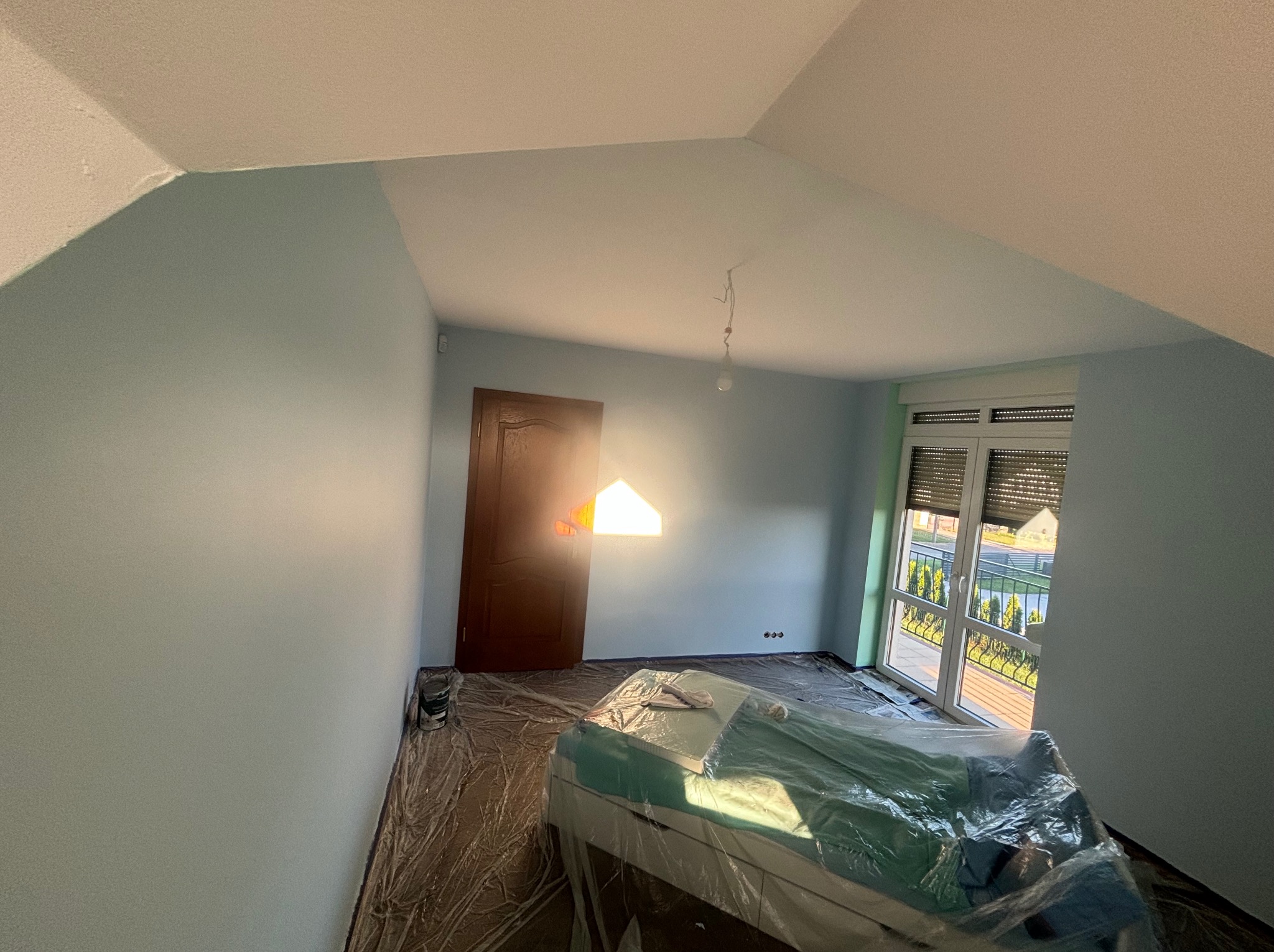 Pokój na poddaszu w trakcie odświeżania, ściany pomalowane na jasny błękit, podłoga zabezpieczona folią malarską, meble przykryte folią ochronną, widoczne puszki z farbą przy ścianie.