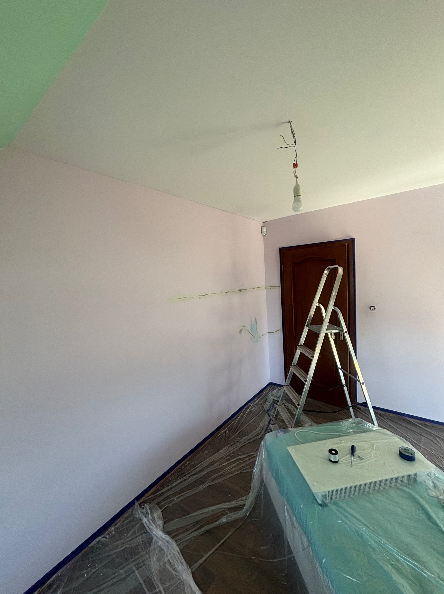 Pokój w trakcie odświeżania: ściany w odcieniach różu i zieleni, zabezpieczona folią podłoga, drabina oparta o obramowane drzwi, grzejnik owinięty folią z narzędziami na wierzchu, widoczne kable...