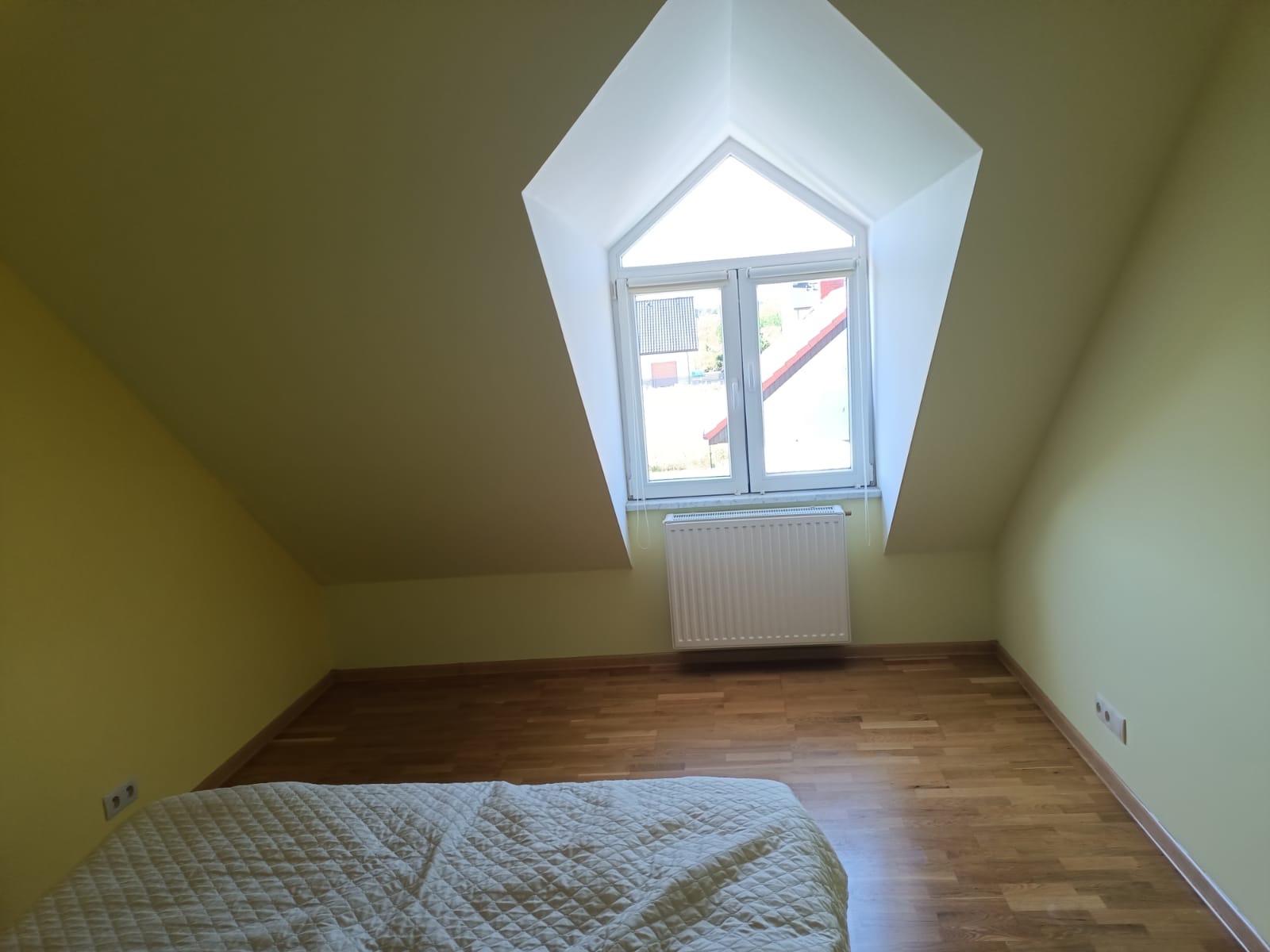 Pokój na poddaszu z oknem w kształcie trapezu, świeżo pomalowane ściany w odcieniach żółci, parkiet ułożony w jodełkę, widoczny fragment łóżka z pikowaną narzutą.