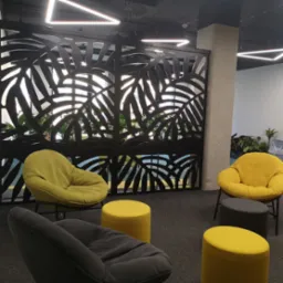 Nowoczesna przestrzeń biurowa z dekoracyjną, ażurową ścianką działową o motywie liści, żółtymi i szarymi fotelami oraz geometrycznym oświetleniem sufitowym.