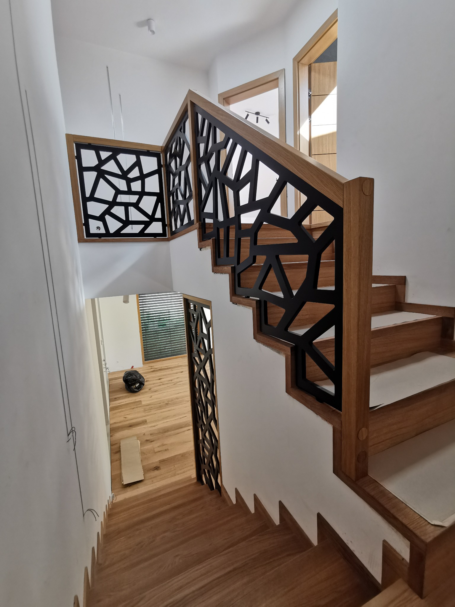 Nowoczesne schody z jasnego drewna z geometryczną, czarną balustradą, widok z góry na dół, z widocznymi elementami wykończeniowymi i podłogą w tle.