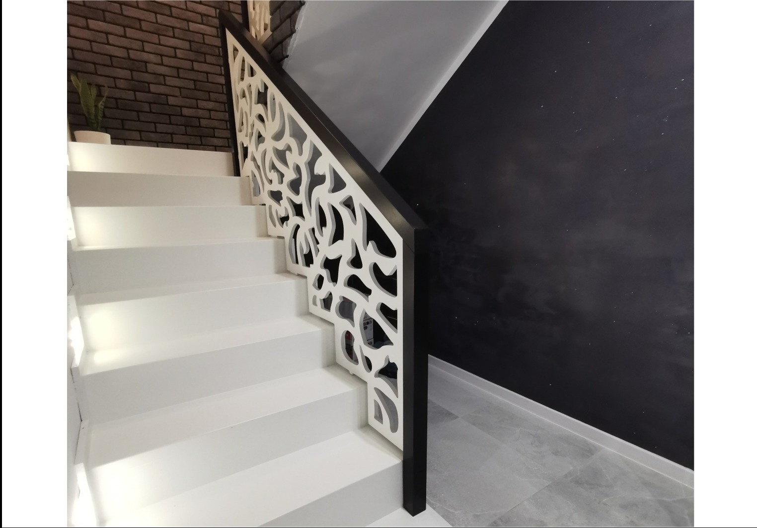 Białe schody z ażurową balustradą o organicznym wzorze, czarny element poręczy, ściana w kolorze grafitowym, fragment ceglanej ściany i rośliny w tle.