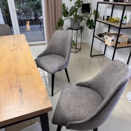 Oskar Pawlak - Nowoczesne, szare krzesła tapicerowane z czarnymi nogami przy drewnianym stole. W tle widoczna roślina Monstera i regał z dekoracjami.
