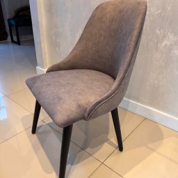 Oskar Pawlak - Tapicerowane krzesło w odcieniach szarości z czarnymi nogami, ustawione na jasnych płytkach. Minimalistyczny design, widoczne detale wykończenia i faktura tapicerki.