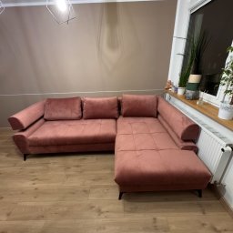 Oskar Pawlak - Różowa, welurowa sofa narożna z niskimi nóżkami na drewnianej podłodze. W tle okno z roślinami i nowoczesna lampa wisząca.