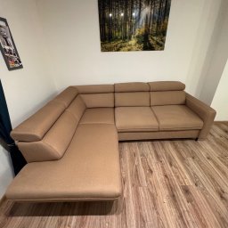 Oskar Pawlak - Beżowy narożnik z regulowanymi zagłówkami na drewnianej podłodze, w rogu pokoju z obrazem lasu na ścianie. Widok z przodu, oświetlenie naturalne.