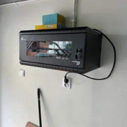 Czarna szafa rack z przeszklonymi drzwiami, zamontowana na ścianie, z widocznymi kablami i urządzeniami wewnątrz, zabezpieczona zamkiem na klucz. Nad szafą leżą pudełka, a obok widoczna instalacja...