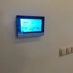 Zamontowany na ścianie panel sterowania systemem alarmowym Hikvision z wyświetlaczem dotykowym, pokazujący aktualną godzinę i datę oraz ikony dostępu do różnych funkcji systemu.