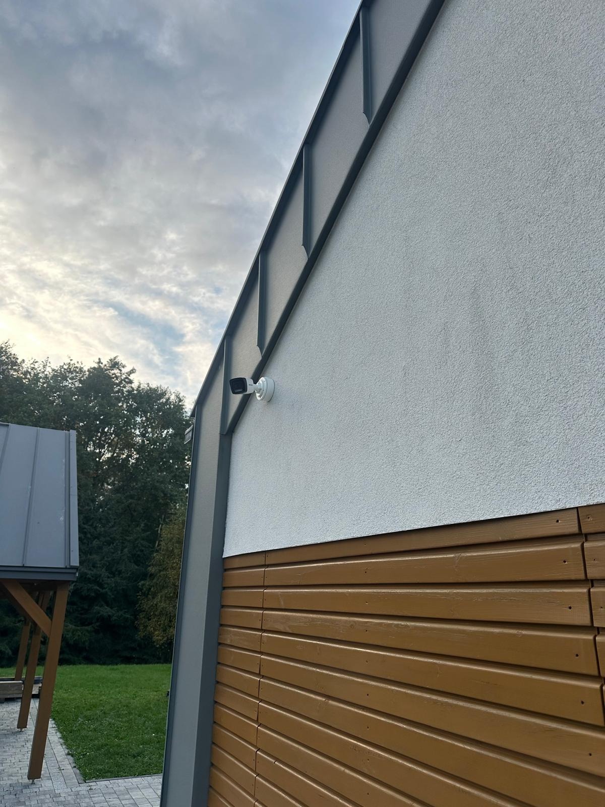 Kamera monitoringu zamontowana na elewacji domu z drewnianą boazerią i szarym dachem, widok z dołu na pochmurne niebo.