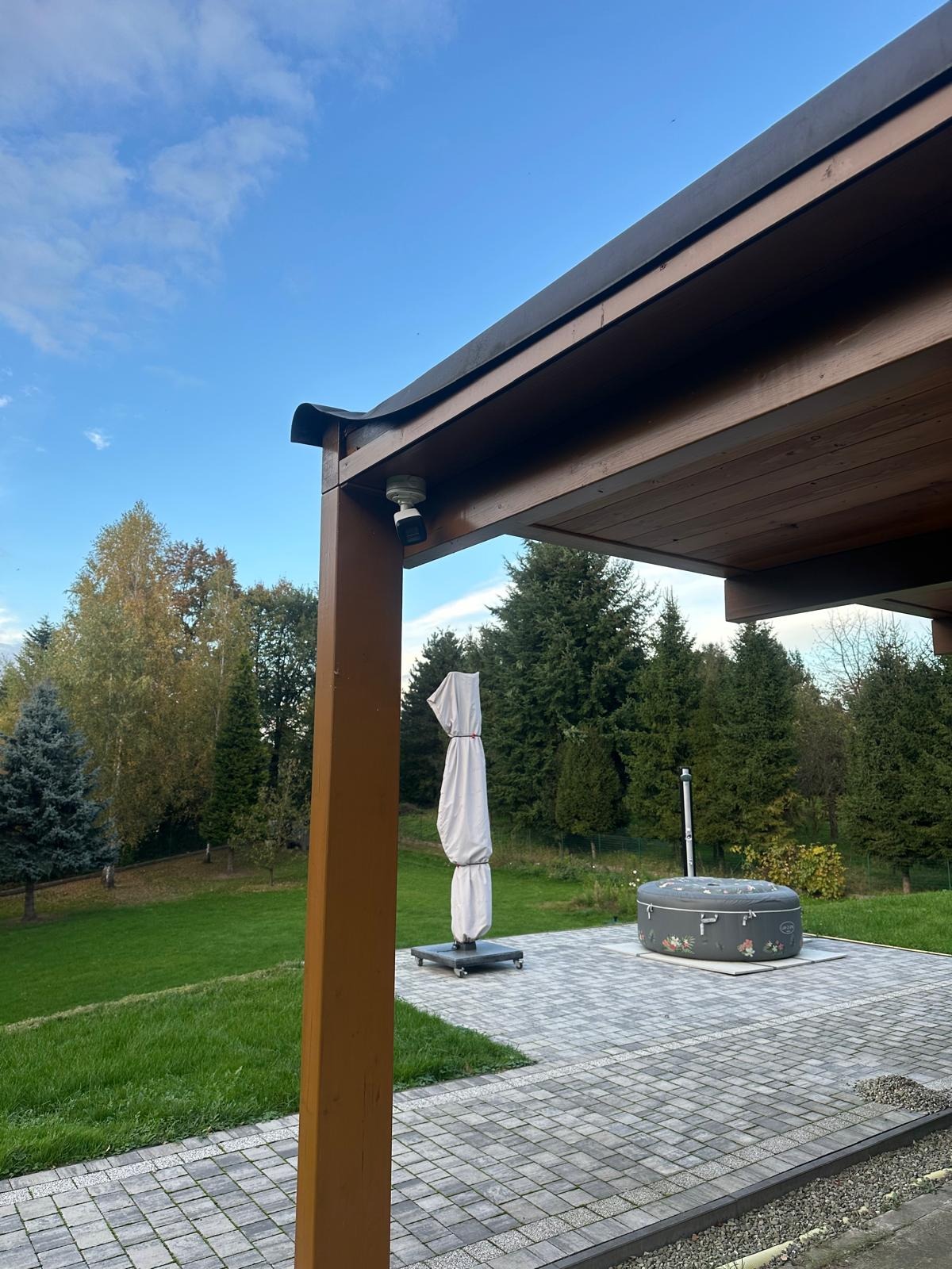 Zewnętrzny monitoring: kamera pod drewnianą pergolą, widok na ogród z jacuzzi i złożonym parasolem.