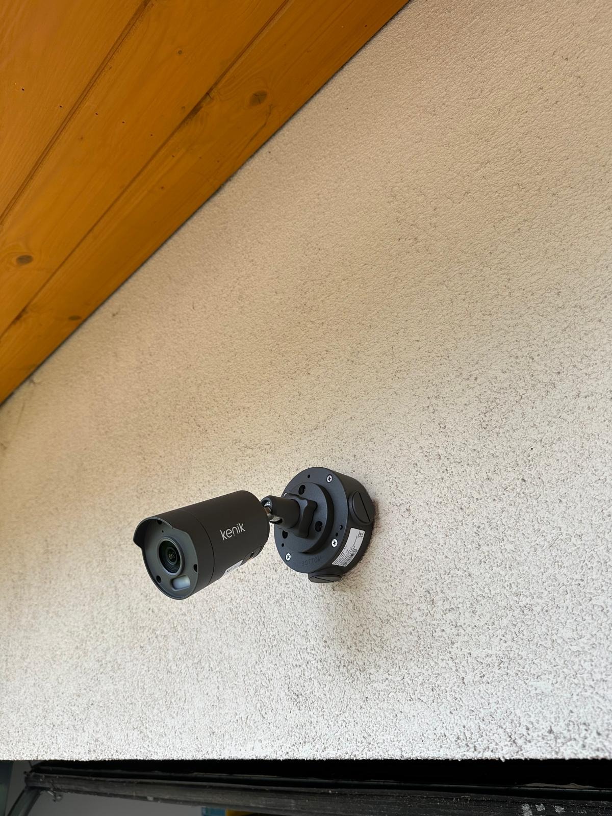 Czarna kamera monitoringu Kenik zamontowana na szorstkiej, beżowej ścianie pod drewnianym okapem dachu.