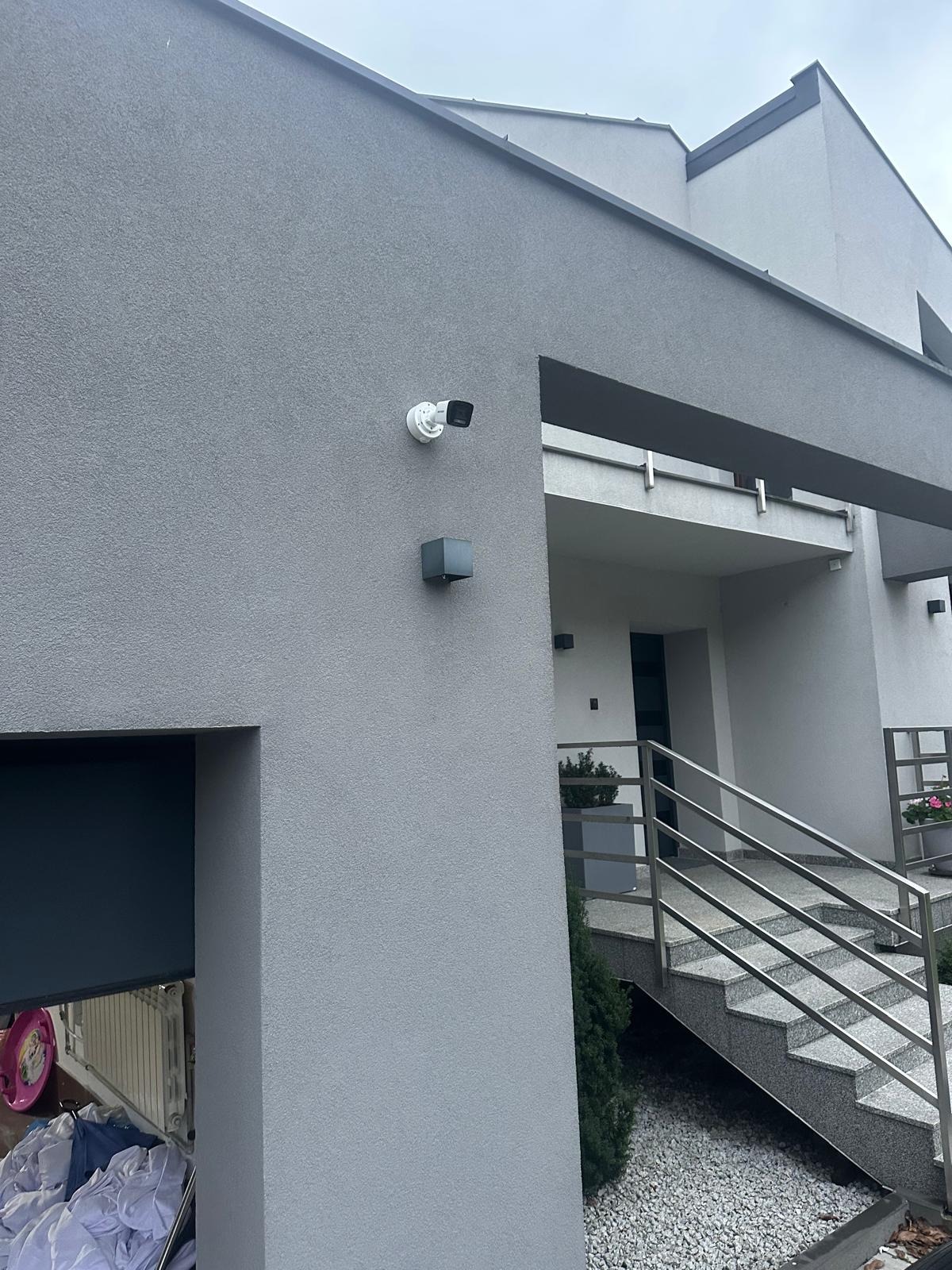 Kamera monitoringu zamontowana na elewacji nowoczesnego, szarego domu jednorodzinnego z metalowymi schodami wejściowymi.