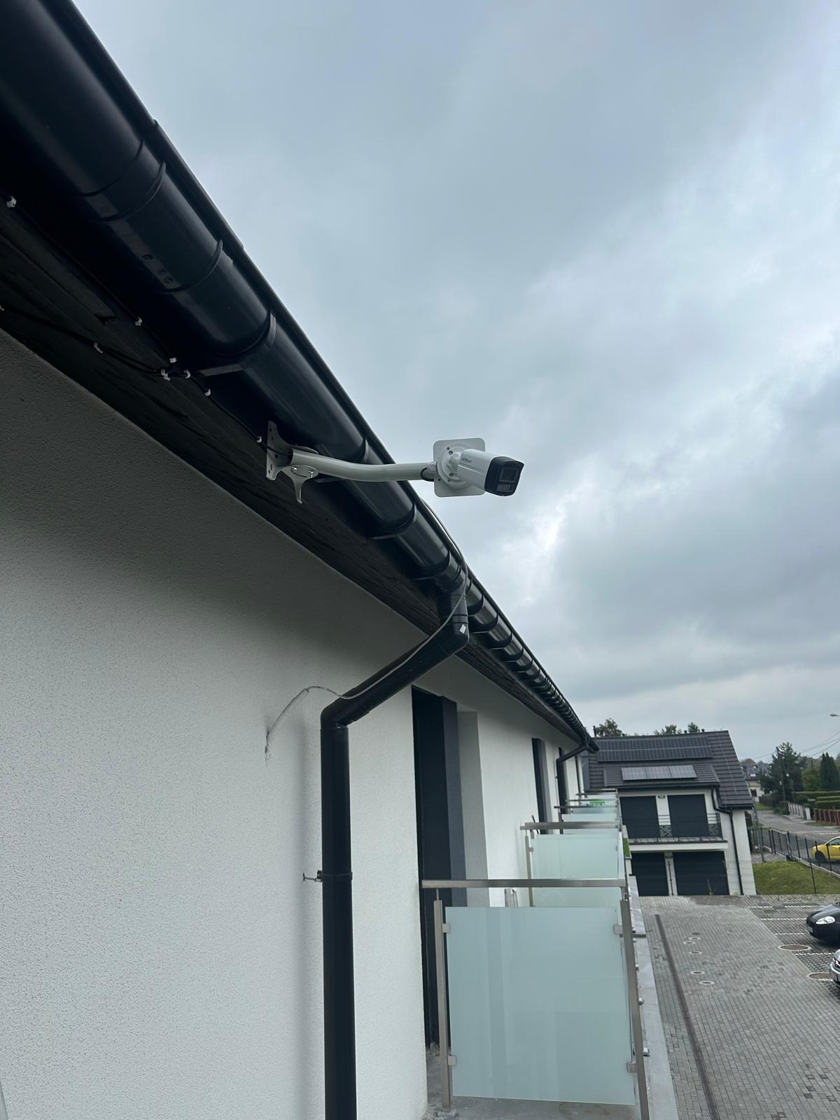 Biała kamera monitoringu zamontowana pod okapem nowoczesnego domu z białą elewacją i czarnymi elementami wykończeniowymi, widok na sąsiedni budynek z panelami słonecznymi na dachu, pochmurne niebo.