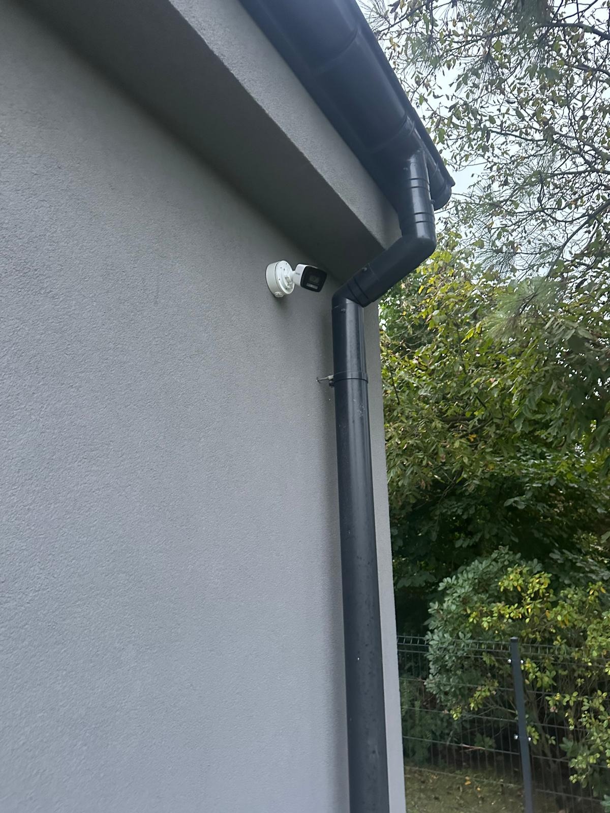 Zewnętrzna kamera monitoringu zamontowana na szarej ścianie domu obok czarnej rynny spustowej, widok z dołu.