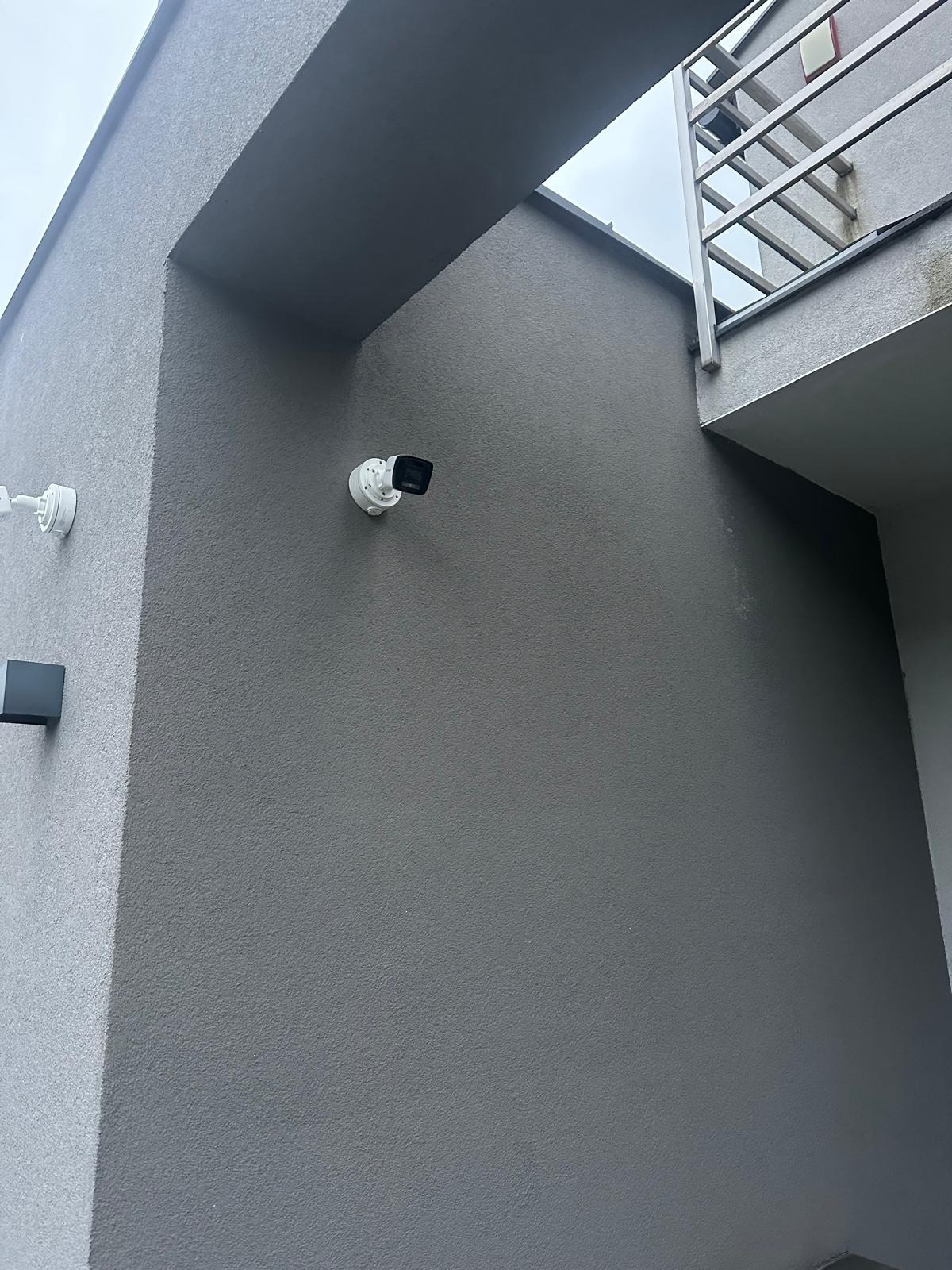 Kamera monitoringu zamontowana na szarej ścianie zewnętrznej budynku z widocznym fragmentem balkonu i oświetleniem.