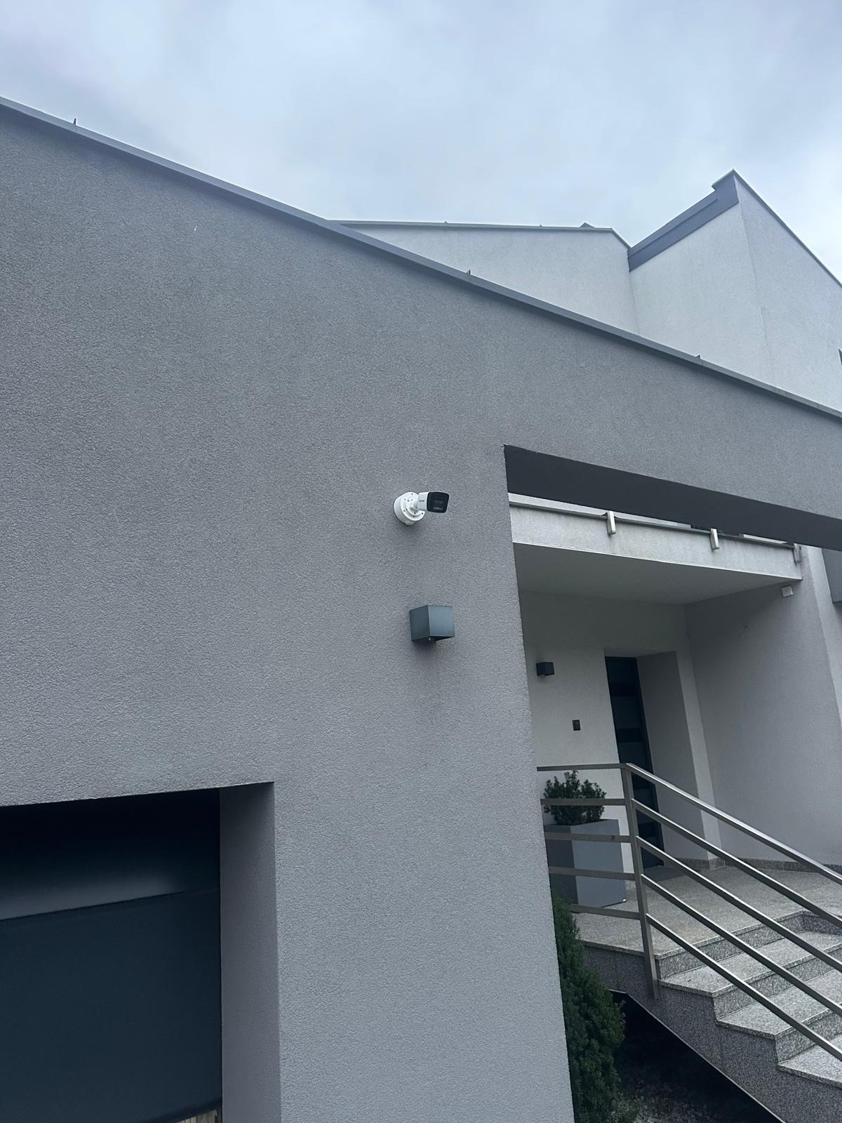 Zewnętrzna kamera monitoringu zamontowana na szarej elewacji nowoczesnego domu jednorodzinnego, widoczny fragment schodów wejściowych z metalową balustradą.