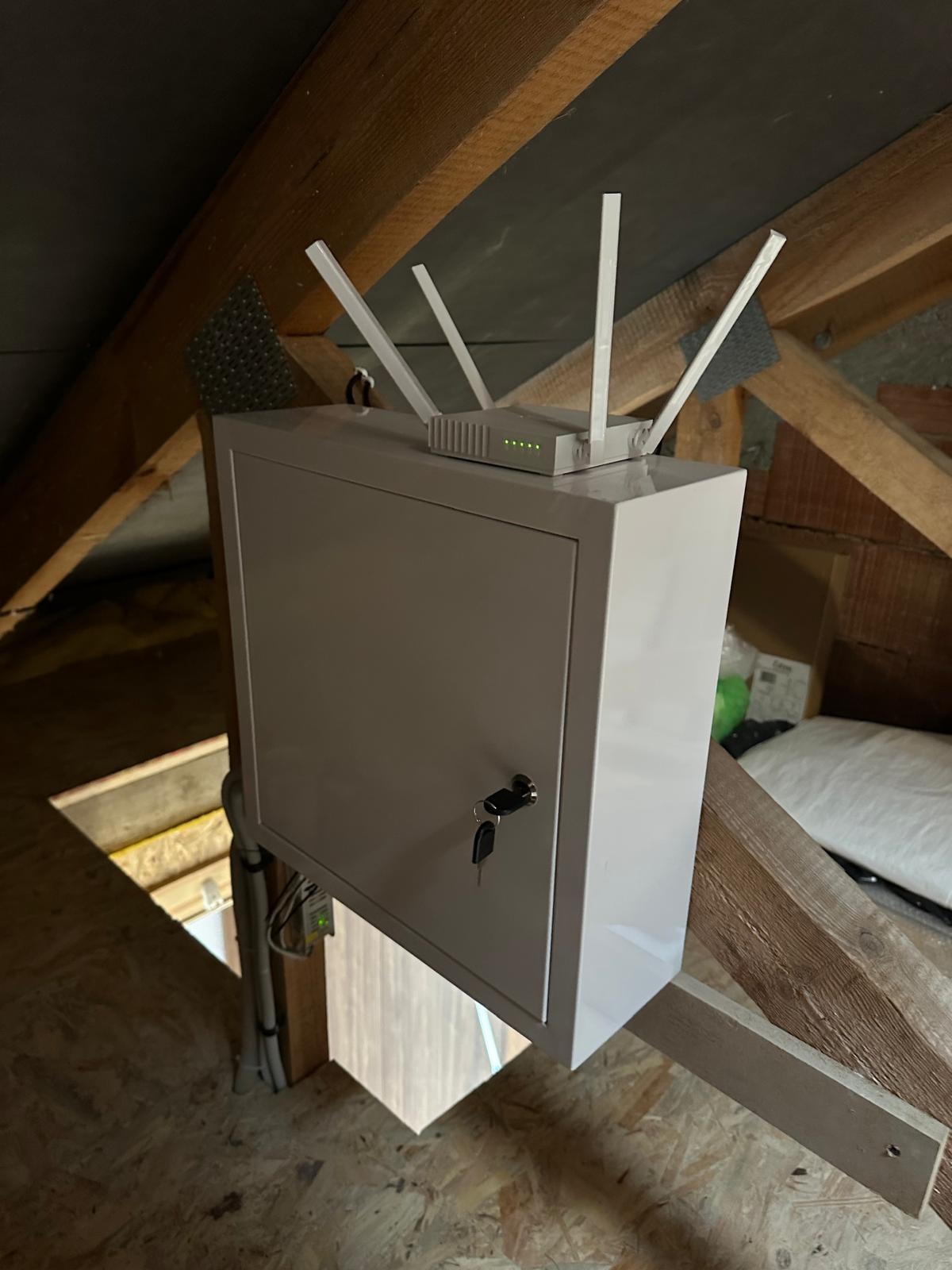 Biała szafka z routerem Wi-Fi na poddaszu, widoczne anteny i diody sygnalizacyjne, kluczyk w zamku, drewniana konstrukcja dachu.