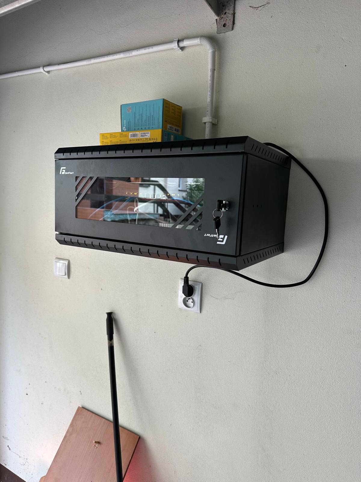 Czarna szafa rack z przeszklonymi drzwiami, zamontowana na ścianie, z widocznymi kablami i urządzeniami wewnątrz, zabezpieczona zamkiem na klucz. Nad szafą leżą pudełka, a obok widoczna instalacja...