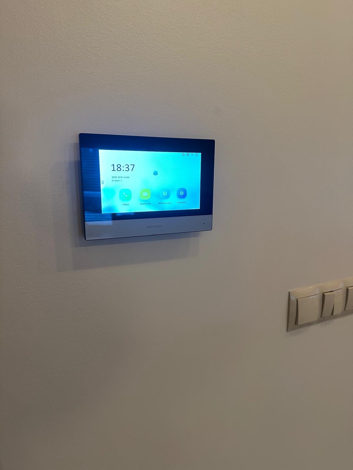Zamontowany na ścianie panel sterowania systemem alarmowym Hikvision z wyświetlaczem dotykowym, pokazujący aktualną godzinę i datę oraz ikony dostępu do różnych funkcji systemu.