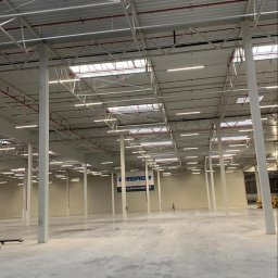 Usługi Elektryczne Adam Jabłoński - Wnętrze hali magazynowej z widoczną konstrukcją dachu, oświetleniem liniowym i instalacją przeciwpożarową w kolorze czerwonym.