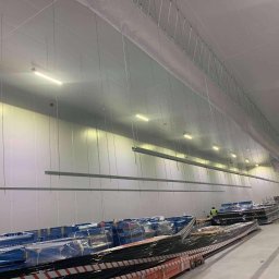 Usługi Elektryczne Adam Jabłoński - Wnętrze hali magazynowej z zamontowanym systemem podwieszeń sufitowych i oświetleniem liniowym LED, widoczne pakiety materiałów budowlanych i pracownik w kamizelce odblaskowej.