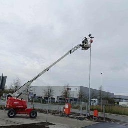 Usługi Elektryczne Adam Jabłoński - Pracownik na podnośniku koszowym montuje oświetlenie na wysokim słupie, widoczne reflektory, tło stanowi budynek przemysłowy z napisem Henne.