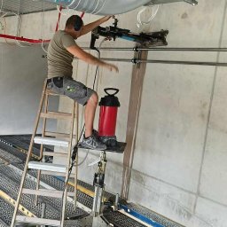 Usługi Elektryczne Adam Jabłoński - Pracownik w słuchawkach montuje system wentylacyjny pod sufitem hali przemysłowej, używając specjalistycznego narzędzia pneumatycznego, stojąc na drabinie z platformą na tle surowych ścian...