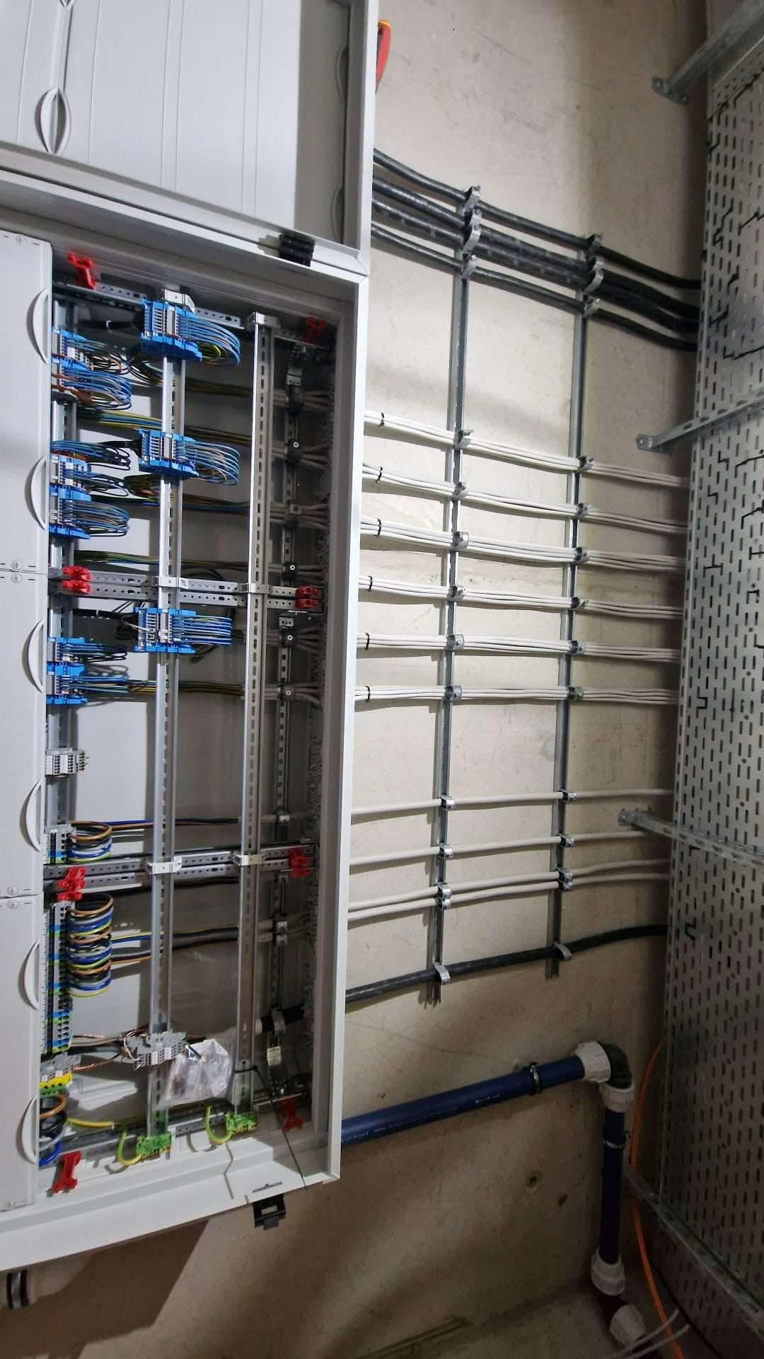 Otwarta skrzynka rozdzielcza z licznymi przewodami i złączkami, kable prowadzone po ścianie w metalowych uchwytach, widoczny fragment instalacji kanalizacyjnej z niebieskiej rury.