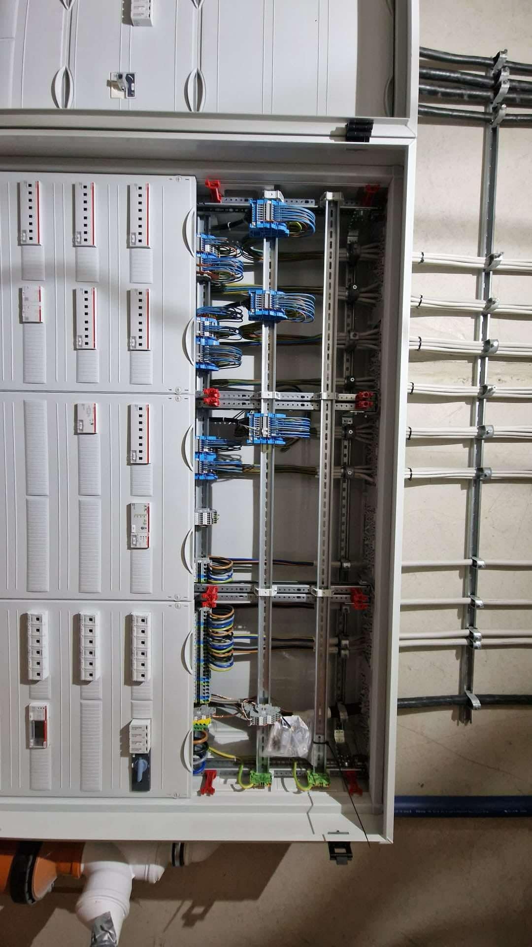 Otwarta skrzynka rozdzielcza z widocznymi bezpiecznikami, przewodami i listwami montażowymi; kable ułożone w równoległych rzędach.