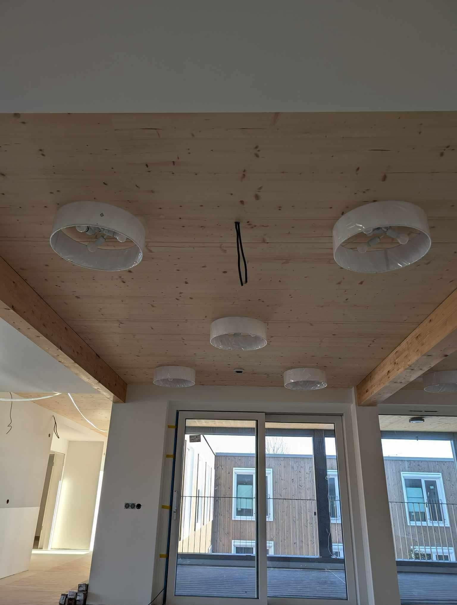 Drewniany sufit z zamontowanymi, owiniętymi folią lampami sufitowymi, widoczne kable i elementy konstrukcyjne, wnętrze w trakcie wykańczania.