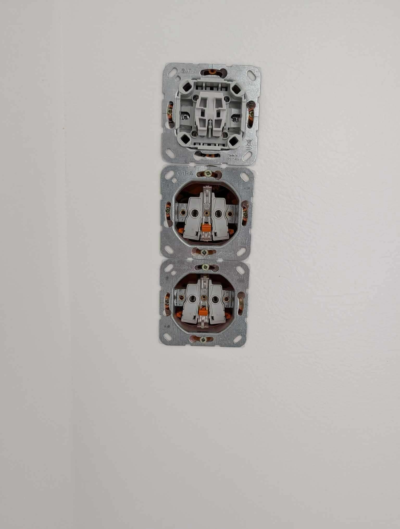 Trzy puszki elektryczne GIRA zamontowane pionowo jedna nad drugą w białej ścianie, widoczne wewnętrzne elementy mocujące i złącza.