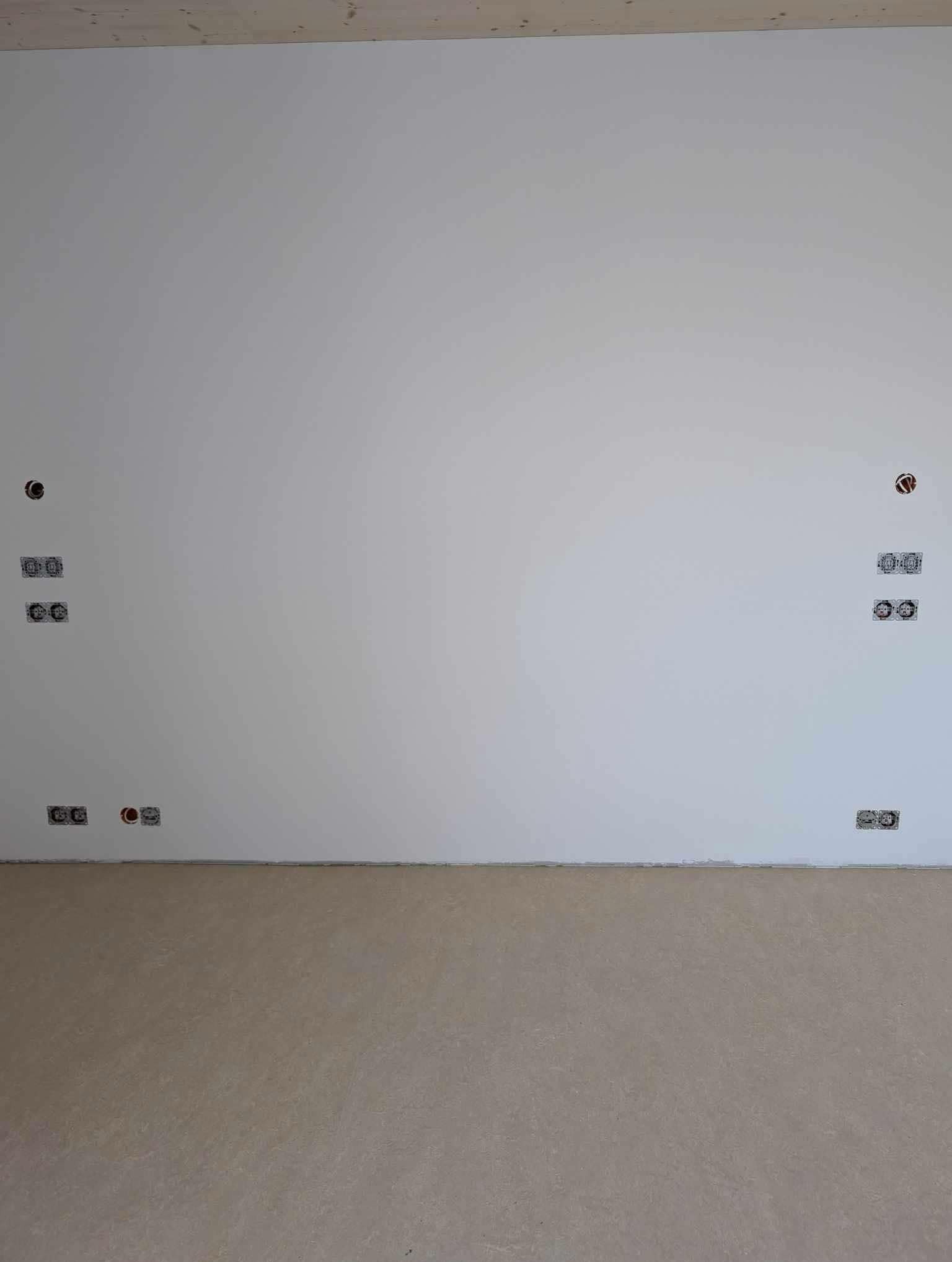 Biała ściana z rozmieszczonymi symetrycznie po obu stronach otworami na gniazdka i puszki elektryczne, widoczne przewody wewnątrz, surowy stan wykończenia wnętrza.