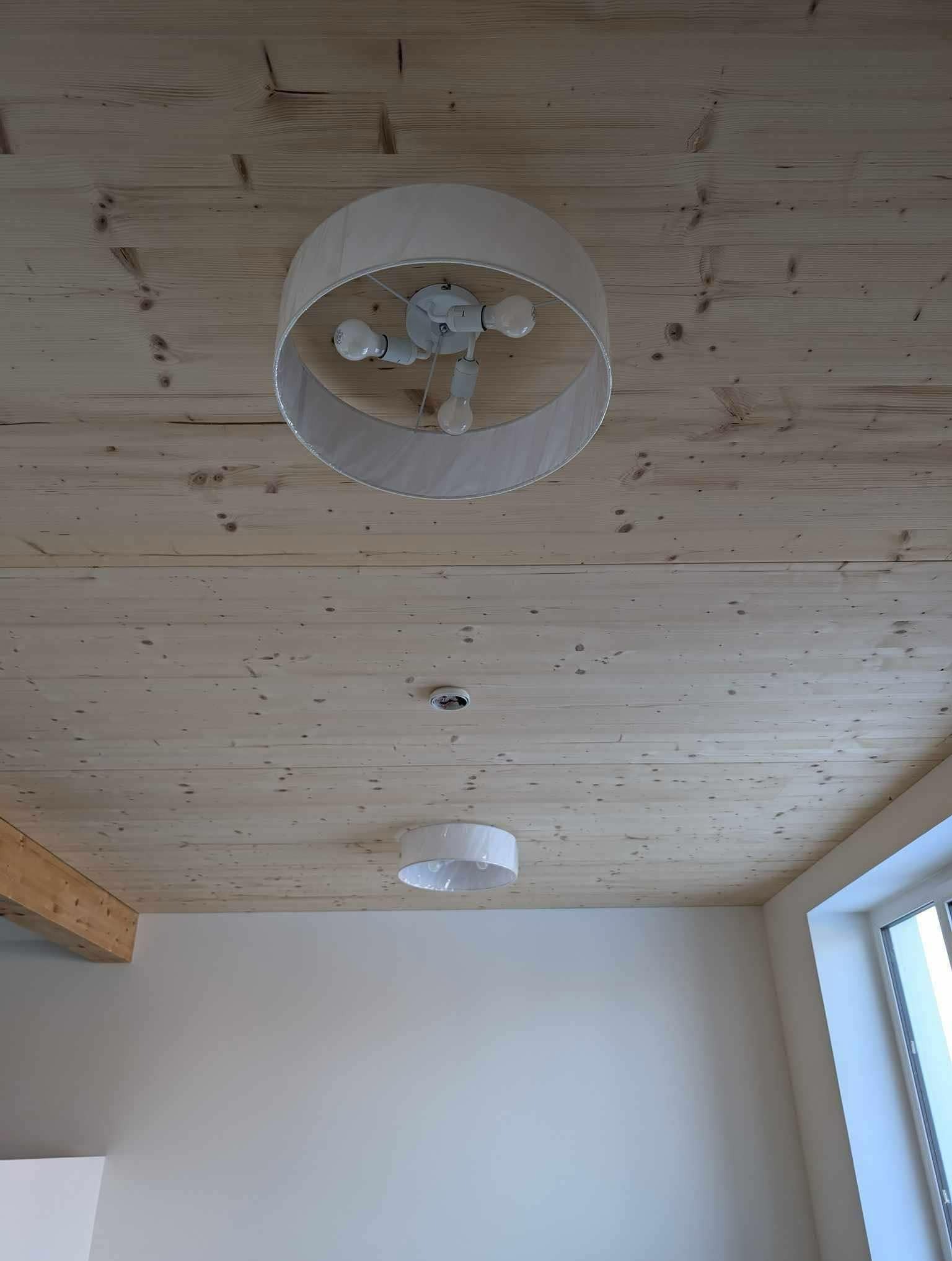 Dwa białe, okrągłe plafony zamontowane na drewnianym suficie z widocznymi sękami, jeden z abażurem, drugi bez, w tle fragment ściany i okna.