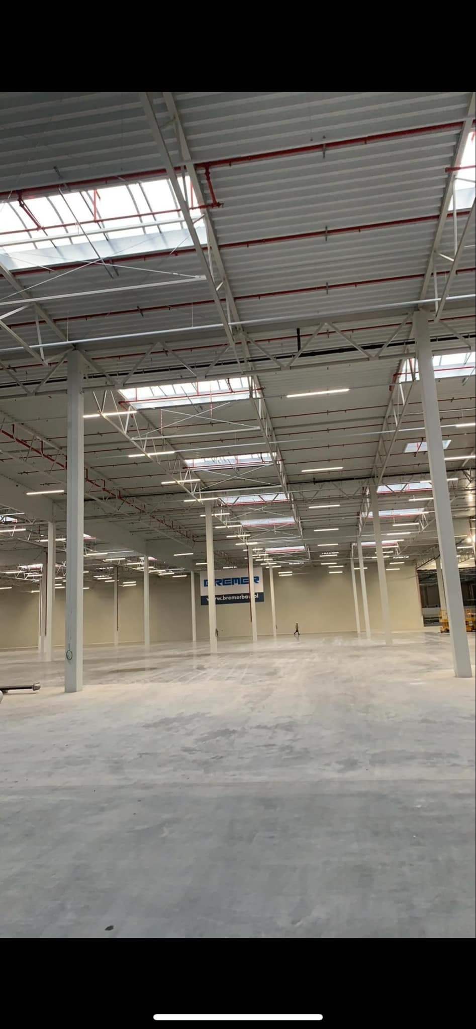 Wnętrze hali magazynowej z widoczną konstrukcją dachu, oświetleniem liniowym i instalacją przeciwpożarową w kolorze czerwonym.