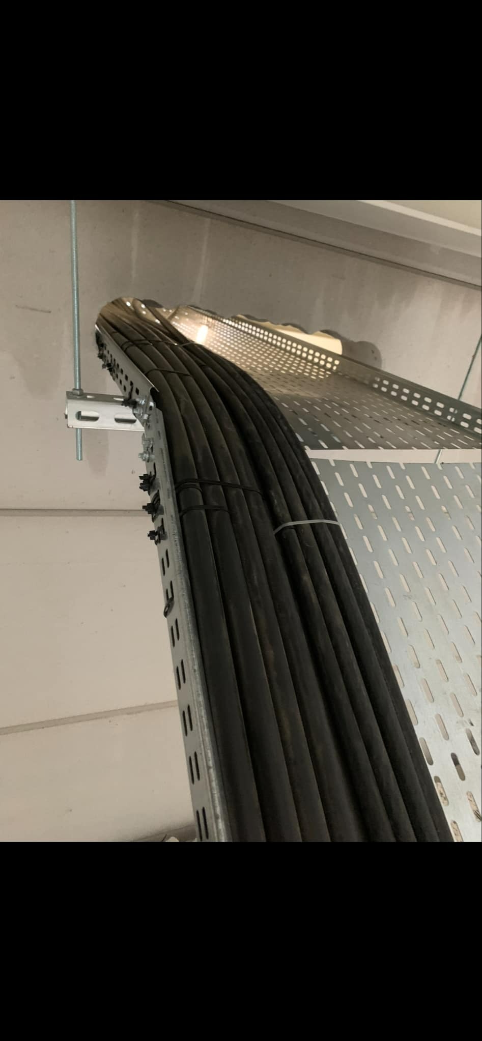 Rząd czarnych kabli elektrycznych, ułożonych w perforowanej metalowej korycie kablowej, zamocowanej do sufitu za pomocą metalowych wsporników i śrub.
