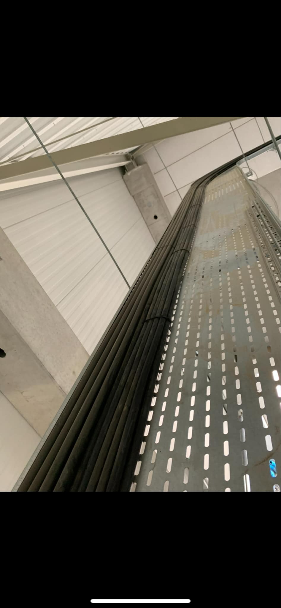 Wiązka kabli elektrycznych ułożona na perforowanej metalowej korycie kablowej, zamocowana pod sufitem w hali przemysłowej.