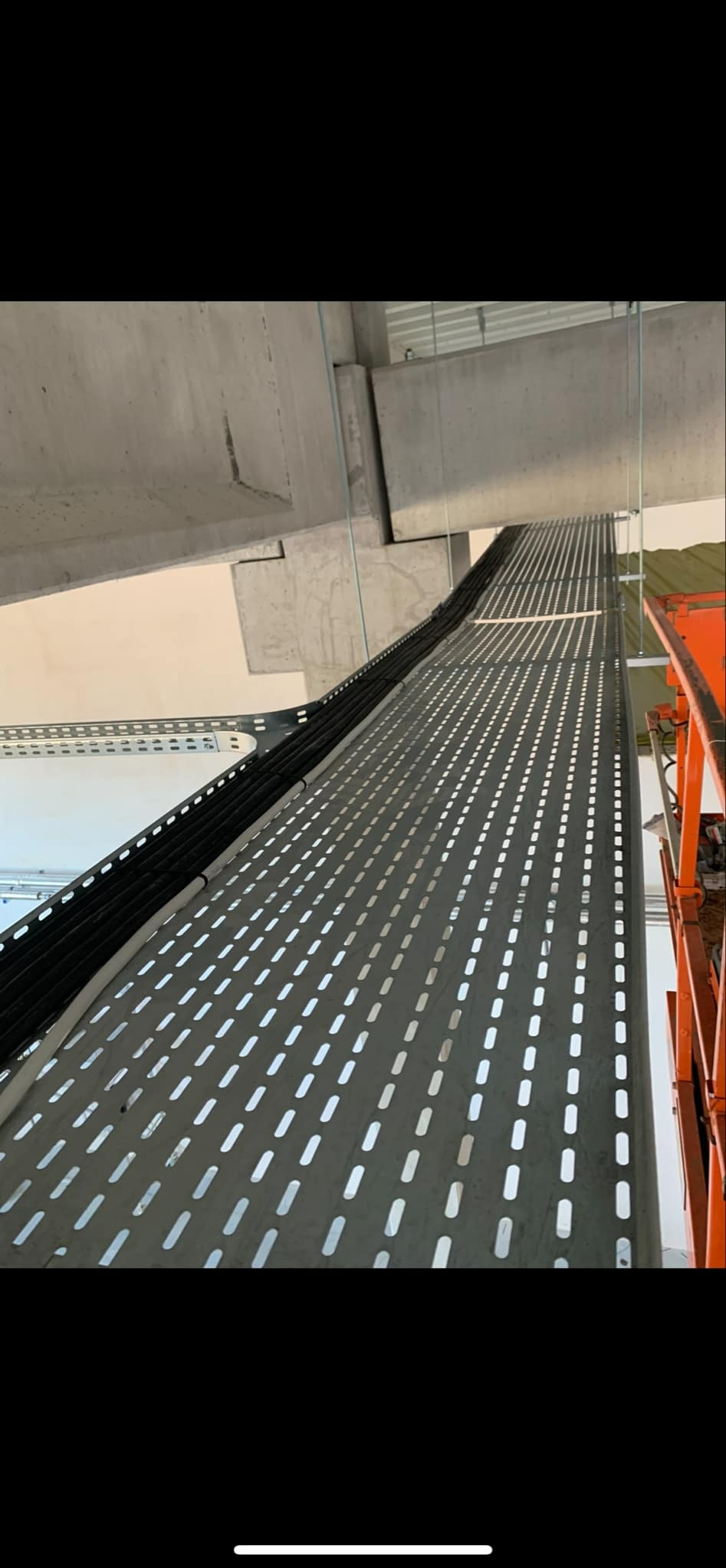 Metalowa korytka kablowa z perforacją, zamocowana pod sufitem, prowadząca czarne kable elektryczne, widok z dołu, fragment podnośnika koszowego po prawej.