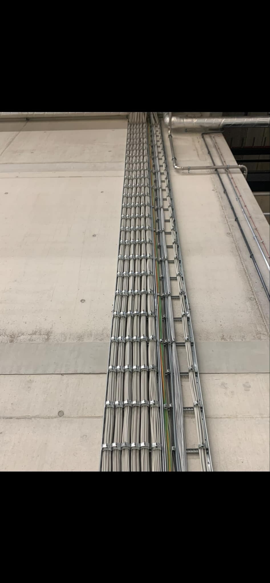 Równolegle ułożone kable elektryczne w metalowych korytkach, zamocowane do betonowej ściany, widoczne rury instalacyjne w tle.